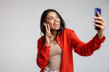 Genç, komik, Afrikalı, sıradan bir bayanın gri üzerine izole edilmiş akıllı telefonuyla özçekim yapması barış jesti göstergesidir.