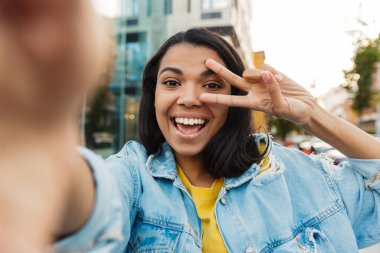 Neşeli Afro-Amerikan kadın görüntüsü barış işareti yapıyor ve şehirde yürürken selfie çekiyor.