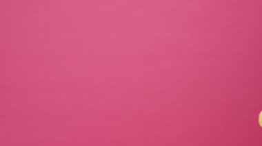 Pembe kazaklı güzel sarışın bir kadın pembe arka planda izole edilmiş bir şekilde yürüyen bir şey ararken elinde sarı bir bavul tutuyor.