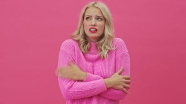 Pembe kazaklı hoşnutsuz sarışın bir kadın donuyor. Pembe bir arka planda tek başına duruyor.