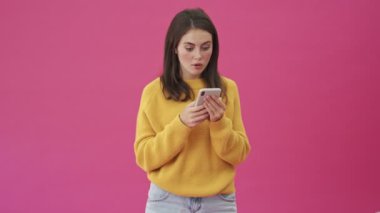 Sarı kazaklı, şaşırmış bir kadın pembe arka planda tek başına duran akıllı telefonuna bakarken birinci sınıf jest yapıyor.