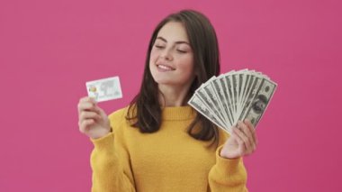 Güzel bir kadın elinde kredi kartı ve para tutuyor. Pembe bir arka planda tek başına duruyor.