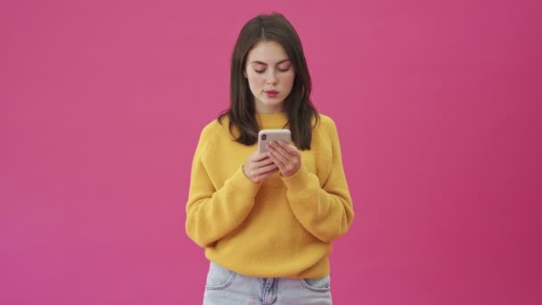 一个穿着黄色毛衣的冷静的女人正在用她的智能手机站在粉色背景上 与外界隔绝 — 图库视频影像
