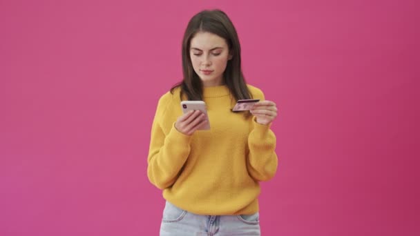一位身穿黄色毛衣 面带微笑的女士一边拿着信用卡一边拿着智能手机 一边站在粉红的背景下独立汇钱 — 图库视频影像