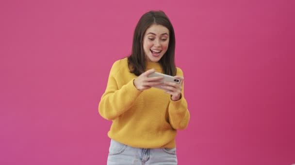 一个穿着黄色毛衣的快乐女人站在粉色背景下 用智能手机玩游戏 — 图库视频影像