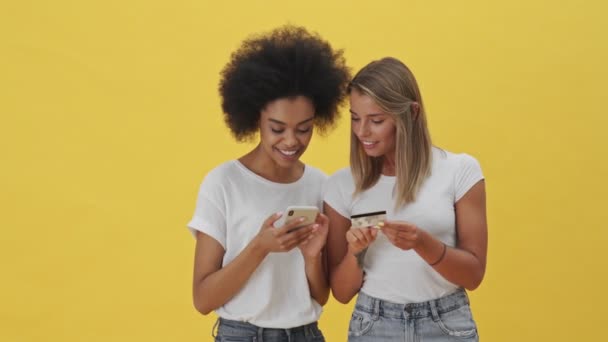 笑顔の女性は黄色の背景に隔離されたクレジットカードスタンドからお金を転送するためにスマートフォンを使用しています — ストック動画