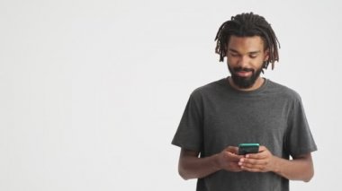 Genç Afrikalı gülümseyen adam beyaz duvar arkasında izole edilmiş cep telefonu kullanıyor ve baş parmağını kaldırıyor.