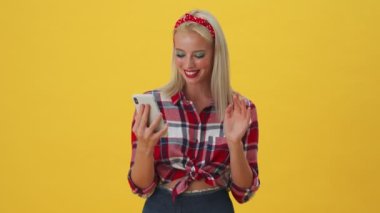Neşeli genç bir kadın, sarı arka planda tek başına duran akıllı telefonunu kullanarak video bağlantısıyla konuşuyor.