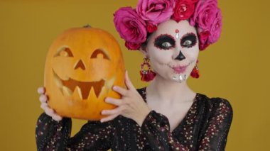 Karnaval kostümü giymiş mutlu bir kadın sarı arka planda izole bir şekilde duran Cadılar Bayramı balkabağını gösteriyor.