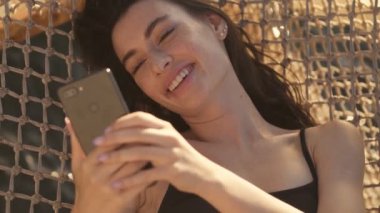 Pozitif genç bir kadın sabah suyun üstündeki hamakta yatarken akıllı telefonunu kullanıyor.