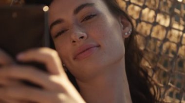 Mutlu, gülümseyen genç bir kadının yakından görüntüsü sabah dışarıda hamakta yatarken akıllı telefonunu kullanıyor.