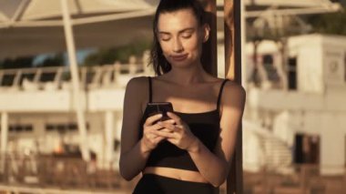 Gülümseyen genç bir kadın sabah sahilde dikilirken akıllı telefonunu kullanıyor.