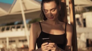 Odaklanmış genç bir kadın sabah sahilde dikilirken akıllı telefonunu kullanıyor.