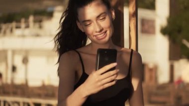 Memnun genç bir kadın sabah kumsalda dururken akıllı telefonunu kullanarak video bağlantısıyla konuşurken elini sallıyor.