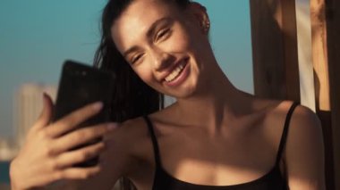 Gülümseyen genç bir kadın sabah kumsalda duran akıllı telefonunu kullanarak video mesajı kaydediyor.