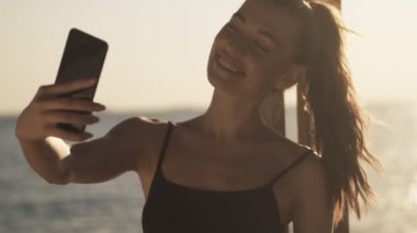 Genç ve güzel bir kadın sabah kumsalda duran akıllı telefonunu kullanarak video mesajı kaydediyor.