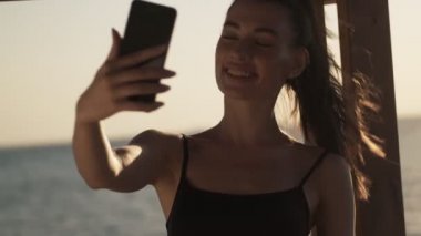 Genç ve güzel bir bayan, sabahları kumsalda duran akıllı telefonunu kullanarak video mesajı kaydediyor.
