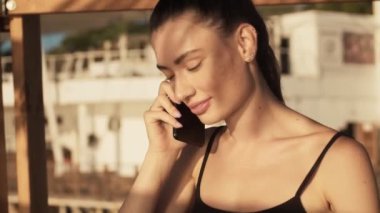 Mutlu, gülümseyen genç bir kadın sabah sahilde akıllı telefonuyla konuşuyor.