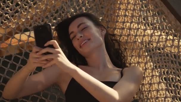 幸せな若い女性は午前中に外のハンモックに横たわっている間 彼女のスマートフォンを使用しています — ストック動画