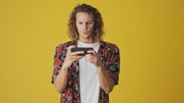 Şok olmuş genç bir adam akıllı telefonuyla bir şey izliyor. Sarı arka planda yatay olarak duruyor.