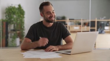 Ofisteki genç mutlu adam dizüstü bilgisayar kullanıyor ve notlar yazıyor.