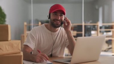 Genç pozitif teslimat yöneticisi ofiste müşteriyle konuşuyor ve paket kutularının yanına notlar yazıyor.