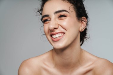 Kıvırcık saçlı, neşeli, yarı çıplak kadın görüntüsü. Gri duvarın diğer tarafına bakıyor ve gülüyor.