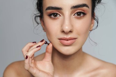 Gri duvardan izole edilmiş kozmetik vitamin kapsülüyle poz veren güzel yarı çıplak kadın görüntüsü.