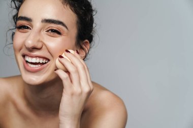 Gri duvarın üzerinden izole edilmiş, gülen ve toz süngeri kullanan neşeli yarı çıplak kadın görüntüsü.