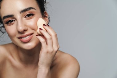 Gri duvarın üzerinden izole edilmiş, gülümseyen ve toz süngeri kullanan yarı çıplak kadın görüntüsü.