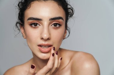 Güzel, yarı çıplak bir kadının, gri duvarda izole edilmiş bir şekilde yüzüne güzellik ürünü sürdüğü görüntüsü.