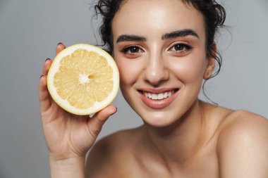 Gri duvarda limonla poz verirken gülümseyen neşeli yarı çıplak kadın görüntüsü