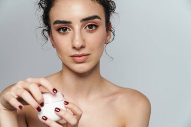 Gri duvardan izole edilmiş kıvırcık saçlı, yüz kremi tutan güzel yarı çıplak bir kadın resmi.