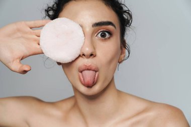 Gri duvarın üzerinde yarı çıplak bir kadının dilini uzatıp pudra süngeriyle dalga geçtiği görüntüsü.