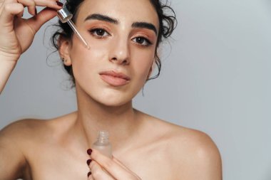 Kıvırcık saçlı, baştan çıkarıcı yarı çıplak kadın görüntüsü gri duvarda izole edilmiş yüz serumu uyguluyor.