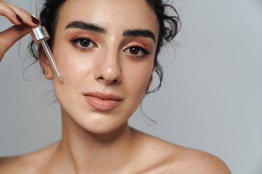 Kıvırcık saçlı, baştan çıkarıcı yarı çıplak kadın görüntüsü gri duvarda izole edilmiş yüz serumu uyguluyor.