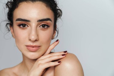 Gri duvarın üzerinden izole edilmiş, kıvırcık saçlı, poz veren ve kameraya bakan güzel yarı çıplak kadın resmi.