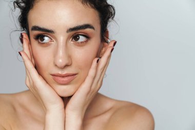 Güzel, yarı çıplak, kıvırcık saçlı bir kadın görüntüsü. Gri duvarın diğer tarafına bakıyor ve poz veriyor.