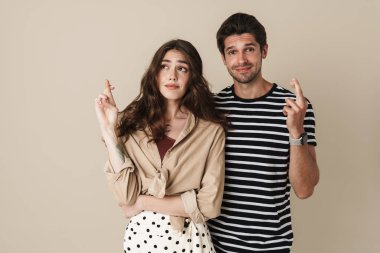 Bej arka planda izole edilmiş, şans dilemek için parmaklarıyla poz veren mutlu bir çift resmi.