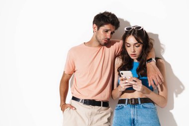 Beyaz arka planda izole edilmiş cep telefonuyla sarılan çekici ve kafası karışık bir çiftin görüntüsü