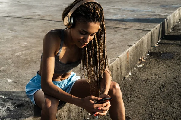 ビーチでリラックスしながら音楽を聴きながらヘッドフォンでスポーツウェアの美しいアフリカの女性 — ストック写真