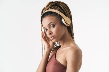 Çekici Afrikalı spor kadını beyaz duvar arkasında izole bir şekilde duruyor, kulaklıkla müzik dinliyor, poz veriyor.
