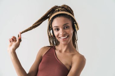 Güzelim gülümseyen Afrikalı esmer kadının portresi Rastalı saçlarıyla beyaz arka planda izole edilmiş kablosuz kulaklıkla müzik dinliyor.