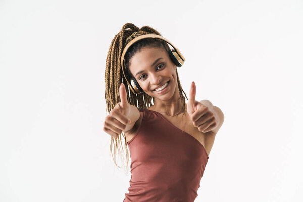 Portrait of beautiful smiling african brunette woman with dreadlocks a listening music via wireless headphones isolated on white background, thumbs up