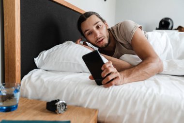 Uykulu genç adam sabahları yatakta yatarken cep telefonu kullanıyor.
