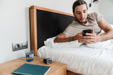 Uykulu genç adam sabahları yatakta yatarken cep telefonu kullanıyor.