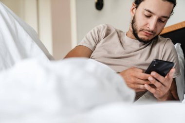 Uykulu genç adam sabahları yatakta yatarken cep telefonu kullanıyor.