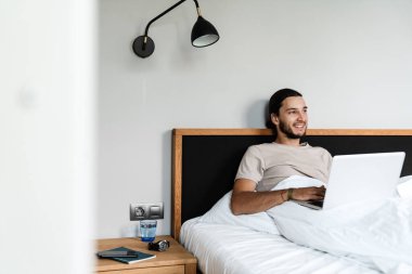 Sabah yatak odasında dizüstü bilgisayarıyla çalışan mutlu bir adam.