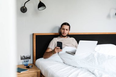 Genç adam sabahları yatarken cep telefonu ve dizüstü bilgisayar kullanıyor.