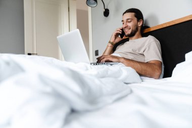 Gülümseyen genç adam sabah yatağında yatarken cep telefonu ve dizüstü bilgisayar kullanıyor.